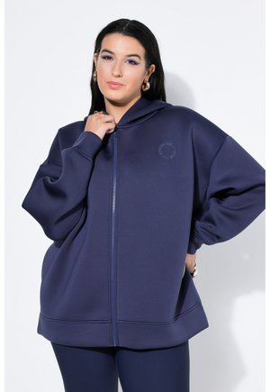 Femme aux longs cheveux foncés portant des boucles d'oreilles créoles dorées et un sweat à capuche zippé bleu marine oversize, debout devant un fond blanc.