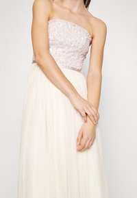 Robe sans bretelles avec un corsage délicat en sequins roses, jupe en tulle crème avec une texture à pois subtils, et ceinture en satin.