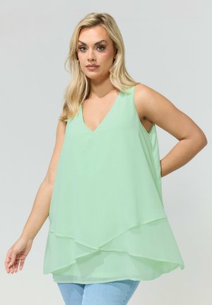 Lyshåret kvinde iført en ærmeløs lagdelt mintgrøn bluse med V-hals og lyseblå jeans, stående foran en ensfarvet baggrund.