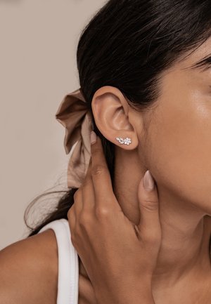 Woman wearing floral-shaped diamond stud earrings, touching her neck, with dark hair tied back by a light brown fabric scrunchie.