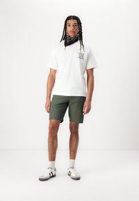 Weißes Baumwoll-T-Shirt mit schwarzem Text auf der Vorderseite, kombiniert mit grünen Shorts, weißen Turnschuhen und einem schwarzen Bandana. Schlichtes Design und Passform.
