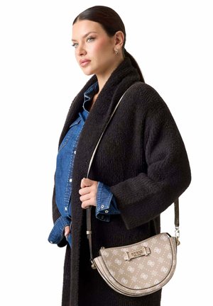 Femme avec une queue de cheval foncée portant un manteau noir texturé et une chemise en jean bleue, portant un sac bandoulière beige à motif demi-lune sur l'épaule.