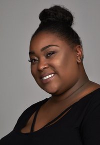 Portret van een persoon met een donkere huid, gekleed in een zwart shirt met een ronde hals. Ze hebben een hoge bun en natuurlijke make-up, terwijl ze glimlachen.