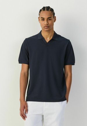 Young man with braided hair wears a black short-sleeve polo shirt and white pants, standing against a plain light gray background.