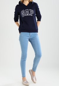 Mörkblå hooded sweatshirt med "GAP" i vita bokstäver, kombinerad med ljusblå skinny jeans och metalliska slip-on skor. Släta tygtextur.