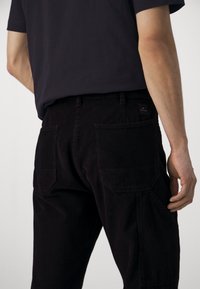 Personne portant un pantalon en velours côtelé noir avec des poches arrière visibles et une chemise noire, montrée de la taille jusqu'en bas sur un fond uni.