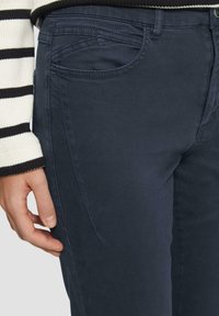 Pantalons en coton bleu marine avec une coupe classique, dotés de poches avant, de coutures subtiles et d'une texture lisse. Main reposant sur le tissu.