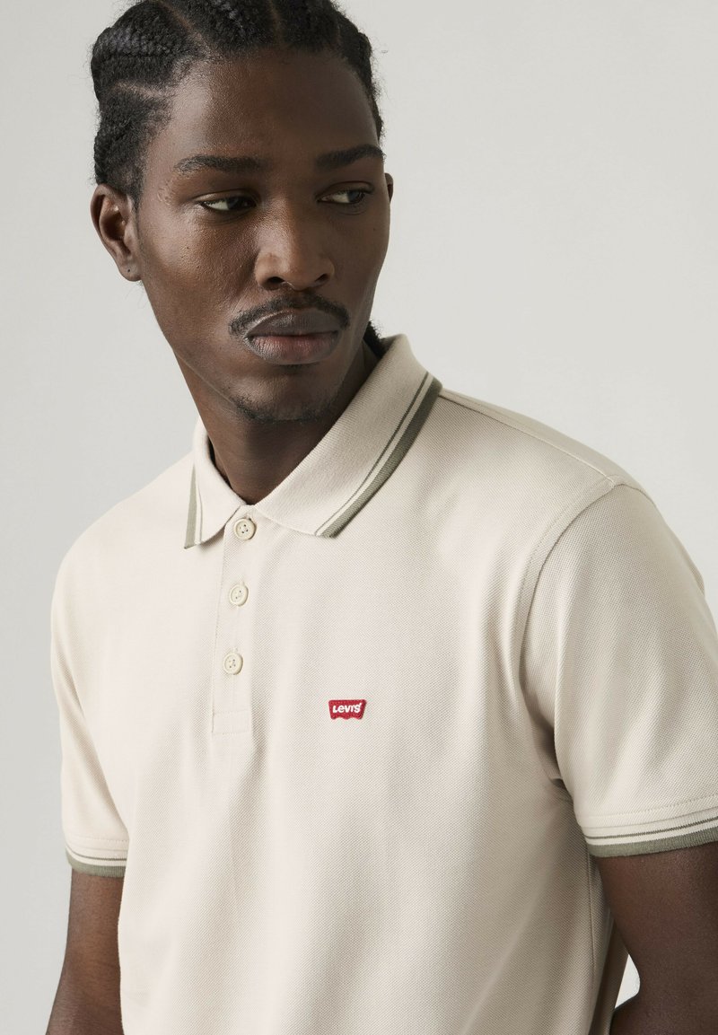 Homme aux cheveux tressés portant un polo beige avec un petit logo rouge Levi's, regardant vers sa gauche sur un fond uni.