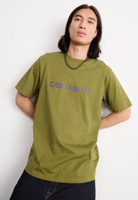 Olivgrünes Baumwoll-T-Shirt mit kurzen Ärmeln, auf der Vorderseite mit einem gedruckten blauen "Carhartt"-Logo. Glatte Textur, lockere Passform.