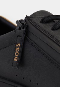 Close-up of black textured sneaker with black laces and zipper pull tab labeled "BOSS" in tan lettering.