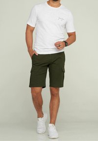T-shirt blanc à manches courtes avec un petit logo et un short cargo vert olive avec des poches latérales, associé à des chaussures de sport blanches.