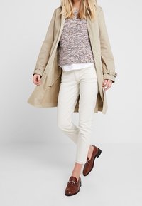 Trench beige sobre un suéter de punto texturizado, camisa blanca y pantalones crema. Los mocasines marrones presentan detalles en plata.