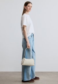 Λευκό βαμβακερό μπλουζάκι συνδυασμένο με ανοιχτό μπλε τζιν ψηλόμεσο. Μια off-white τσάντα με καμπυλωτή λαβή κρατείται στο δεξί χέρι.