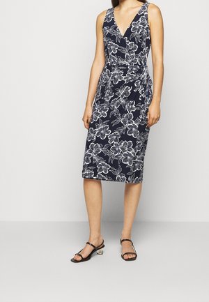Femme portant une robe portefeuille sans manches bleu marine avec imprimé floral blanc et des sandales noires à talons bas, debout devant un fond clair uni.