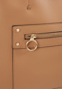 Surface en cuir fauve avec une petite poche zippée équipée d'un anneau coulissant circulaire en or et de détails à rivets dorés.