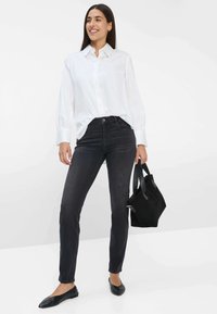 Witte blouse met lange mouwen, zwarte high-waisted jeans en zwarte platte schoenen. Model heeft een kleine zwarte tas bij zich.