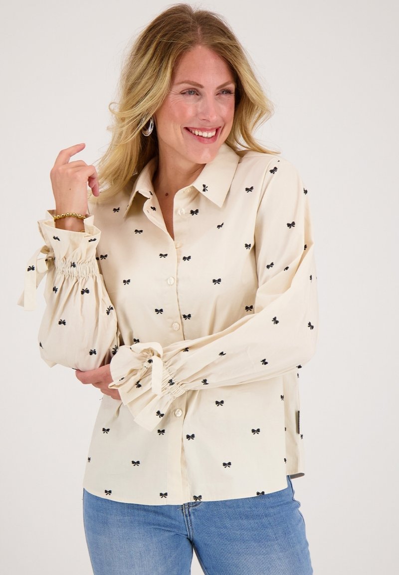 Romantisch crèmekleurig overhemd met een zwart strikpatroon, elastische ballonmouwen met strikdetails en een klassieke kraag. Draagt bij blauwe spijkerbroek.