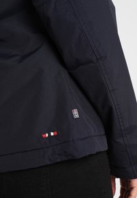 Veste d'hiver bleu marine en tissu lisse et résistant à l'eau, avec une petite étiquette de logo et des détails de couture rouge, blanc et bleu.