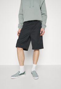 Svarta shorts med en avslappnad passform, bärs med en ljusgrön hoodie och grön-och-vit rutiga slip-on skor. Vita ankelsockor.