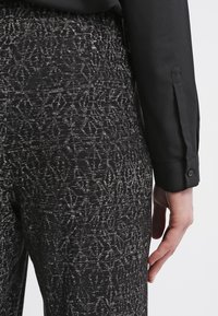 Czarne spodni z teksturą i białym abstrakcyjnym wzorem, połączone z dopasowaną czarną koszulą z zapięciem na guziki przy mankietach. Skupienie na widoku z tyłu.