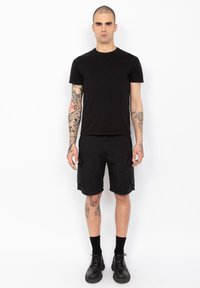 Zwarte katoenen t-shirt gecombineerd met zwarte cargoshorts. Model draagt zwarte sokken en laarzen. Tatoeages zichtbaar op armen en benen.
