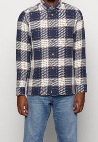 Camicia button-up in fantasia a quadri blu navy, crema e rosso. Realizzata in tessuto testurizzato con colletto e bottoni in legno. Accentuata da una toppa con logo.