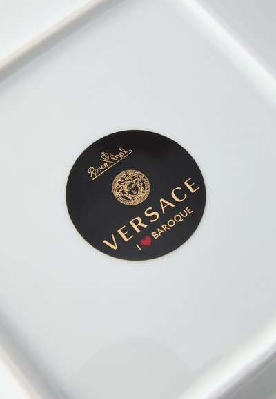 Balta keramikas šķīvis ar apaļu melnu uzlīmi, uz kuras ir zelta teksti un rotājumi, tostarp Medūzas simbols un uzraksts "I ❤️ BAROQUE".