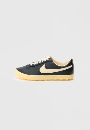 Black low-top Nike sneaker with cream-colored swoosh, laces, and sole on a plain light gray background.