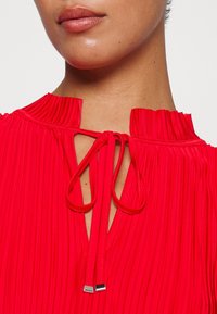 Rød plisseret bluse med høj hals og knyt-lukning. Har en nøglehul-udskæring og tekstureret stof med glatte snore og metaldetaljer.
