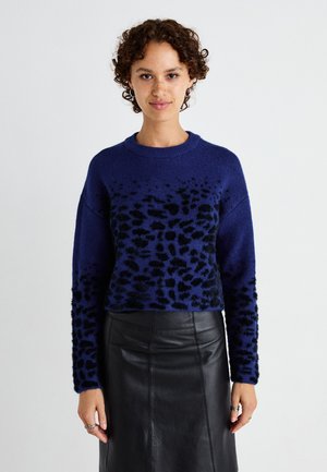 Pull bleu marine avec motif léopard noir, tissu texturé, col rond, manches longues, porté sur une jupe en cuir noir.