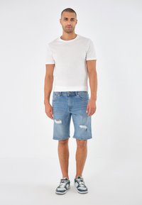 Hvit kortermet t-skjorte kombinert med lyseblå ripped denimshorts og grå joggesko, stående på en ensfarget hvit bakgrunn.