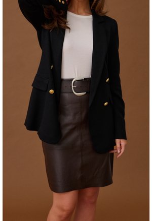 Vrouw draagt een zwarte blazer met gouden knopen, witte top, donkerbruine leren rok en brede donkere riem met grote zilveren gesp.