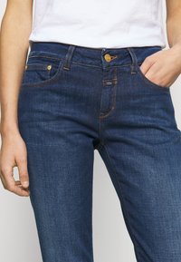 Gros plan sur une personne portant un jean en denim bleu foncé avec des coutures orange et un t-shirt blanc, une main dans la poche, l'autre détendue le long du corps.