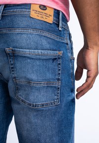 Jeans de denim azul oscuro con un aspecto desgastado, con costuras naranjas, dos bolsillos traseros y una etiqueta de cuero marrón en la cintura.