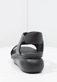 Sandalo in pelle nera con suola arrotondata, schienale aperto e cinturini larghi. Il materiale testurizzato e il design minimalista evidenziano la funzionalità.