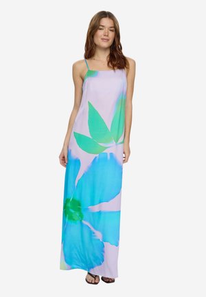 Žena v dlhých šatách bez rukávov s veľkým modro-zeleným kvetinovým vzorom stojaca pred jednoduchým pozadím.