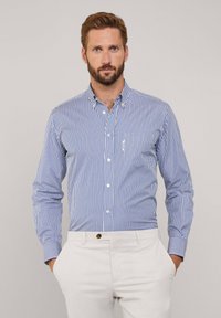 Camicia button-up a righe verticali blu e bianche, maniche lunghe, taschino sul petto, tessuto di cotone. Abbinata a pantaloni di colore chiaro.