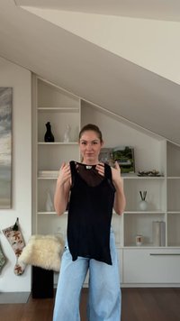 Must varrukateta pluus läbipaistva kaelusega, millel on lõdvestunud lõige ja kõver ääris. Modell esitleb riietust heledas siseruumis.