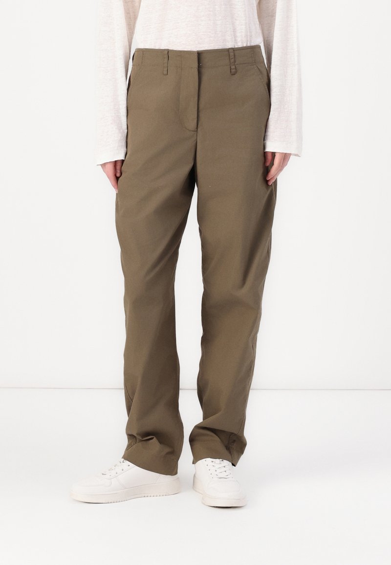 Pantalon kaki à jambes droites en tissu lisse, avec braguette à glissière, passants pour ceinture et poches latérales, porté avec des baskets blanches.