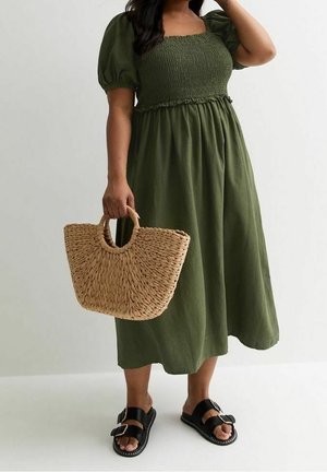 Kvinde iført olivengrøn smockkjole og sorte sandaler med spænde, holder tanfarvet flettet stråtaske med runde håndtag.