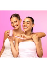 Dwie kobiety pozują na tle w kolorze różowym, każda trzyma różowe produkty do pielęgnacji skóry. Produkty obejmują cylindryczny słoik i aplikator w formie sztyftu.