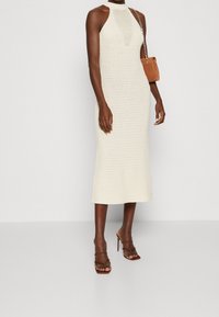 Robe halter tricotée, crème, avec un corsage ajusté et une jupe de longueur mi-mollet. Associée à des talons marron à lanières et un petit sac à bandoulière marron.