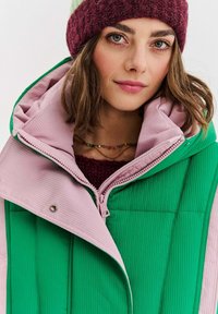 Vrstvená vesta v zelenej a ružovej farbe, s rebrovanou textúrou, veľkým golierom a zapínaním na zips. Pod ňou je vidieť bordový pletený sveter.