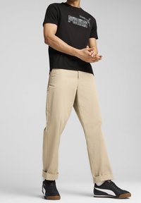 Svart bomulls t-shirt med "PUMA"-logotyp, beiga bomullsshorts, och svarta sportskor med vita detaljer, i stående position med händerna hopknäppta.