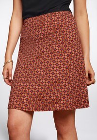Red patterned skirt with geometric design in shades of orange, black, and white. A-line shape, mid-length, with a smooth woven texture.
