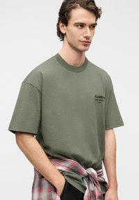 Πράσινο ελιάς βαμβακερό T-shirt με στρογγυλή λαιμόκοψη και κοντά μανίκια· διαθέτει μικρό μαύρο λογότυπο στο στήθος. Χαλαρή εφαρμογή, κρατείται από ένα πουκάμισο καρό.