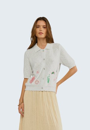 Femme portant un cardigan blanc à manches courtes boutonné avec une broderie sur le thème de la mer et une jupe plissée beige, posant avec une main sur la hanche.