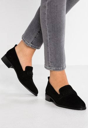 Personne portant un jean gris et des mocassins en daim noir à petit talon, debout sur un sol blanc devant un fond uni.