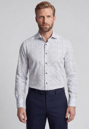 Man draagt een wit overhemd met lange mouwen met een klein donker patroon, gecombineerd met een donkerblauwe broek, staand tegen een effen achtergrond.