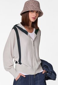 Helle beige Zip-Hoodie mit dunkelgrünen Streifen, aus leichtem Material und mit Kapuze. Kombiniert mit einem schlichten weißen Shirt und einer Denim-Hose.